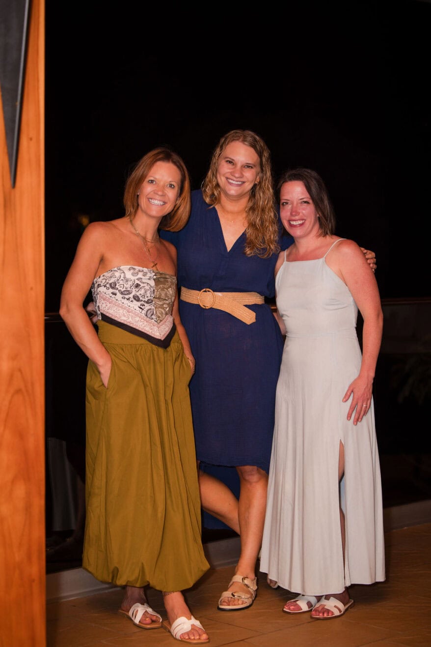 Three women stand together and smile at night, looking like friends celebrating after a custom home build. One wears a strapless top with an olive skirt, another sports a navy dress with a tan belt, and the third is in a light gray sleeveless dress.