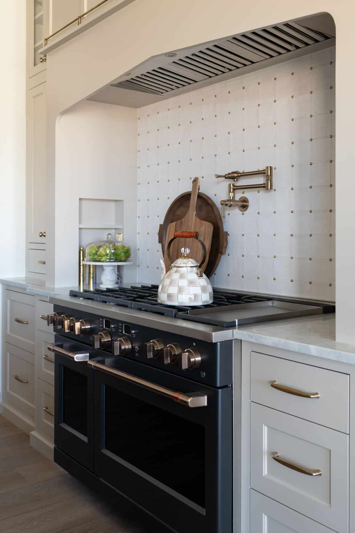 A modern kitchen in a custom home features a black double oven, a white checkered teapot on the stove, marble backsplash, wooden cutting board, stylish decor, and sleek light gray cabinets with gold handles—a perfect touch for your home build.