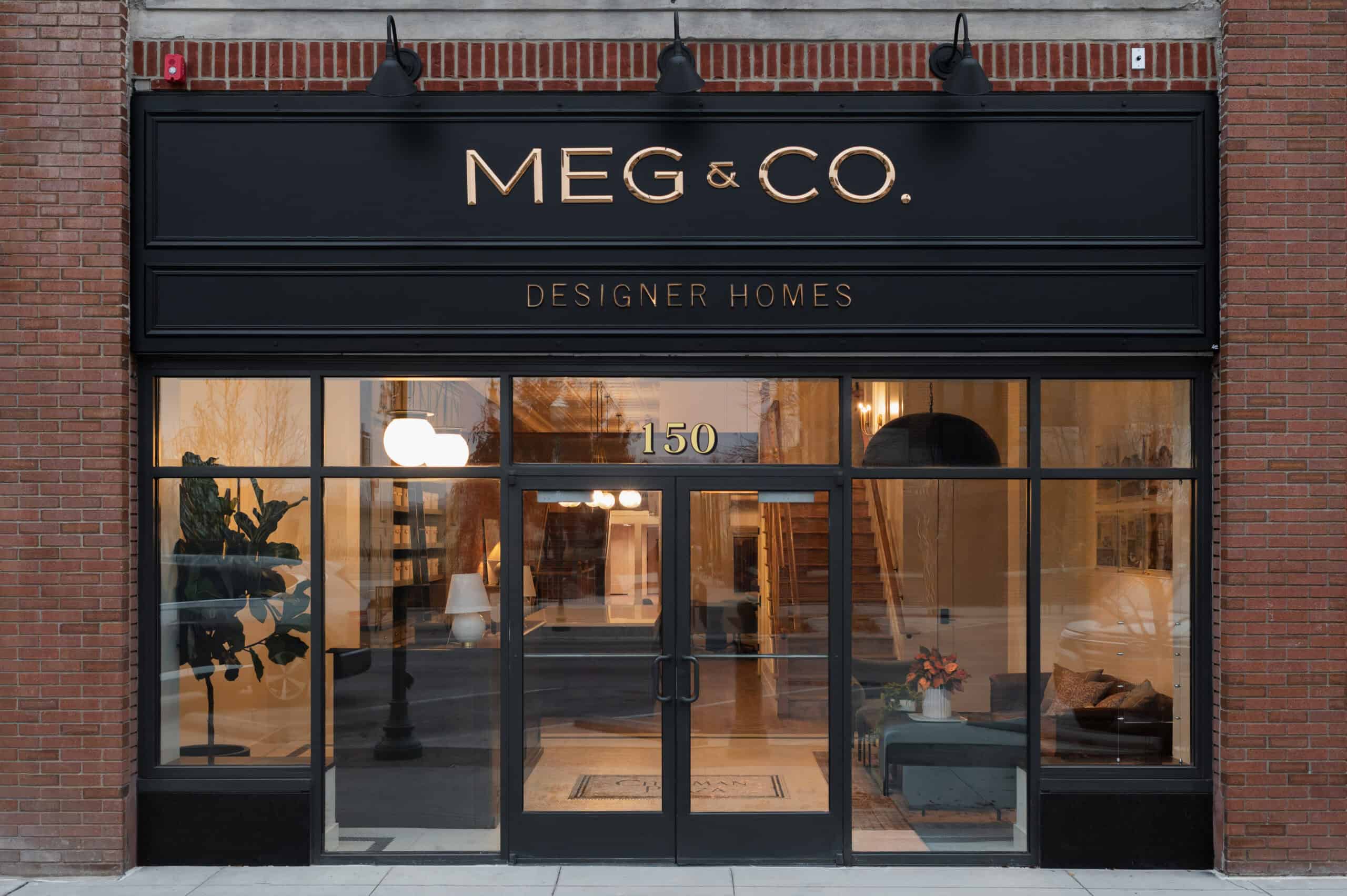 The storefront of MEG & CO. Designer Homes features large glass windows and doors, with modern decor visible inside. The shop’s black sign with gold lettering is displayed above the entrance at 150.