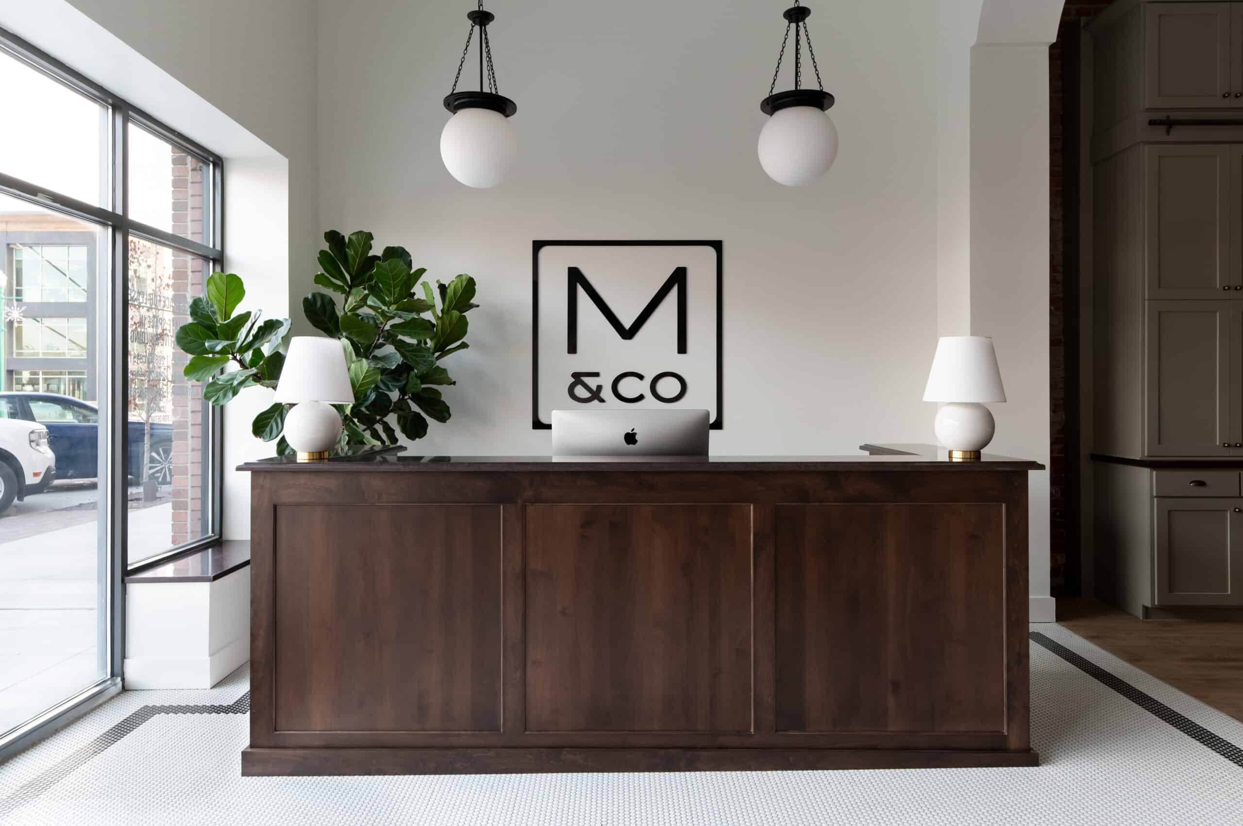 A modern reception area with a dark wood desk, two white lamps, a computer, green plant, and a large M & Co logo on the wall. Two round pendant lights hang from the ceiling. Large windows let in natural light.