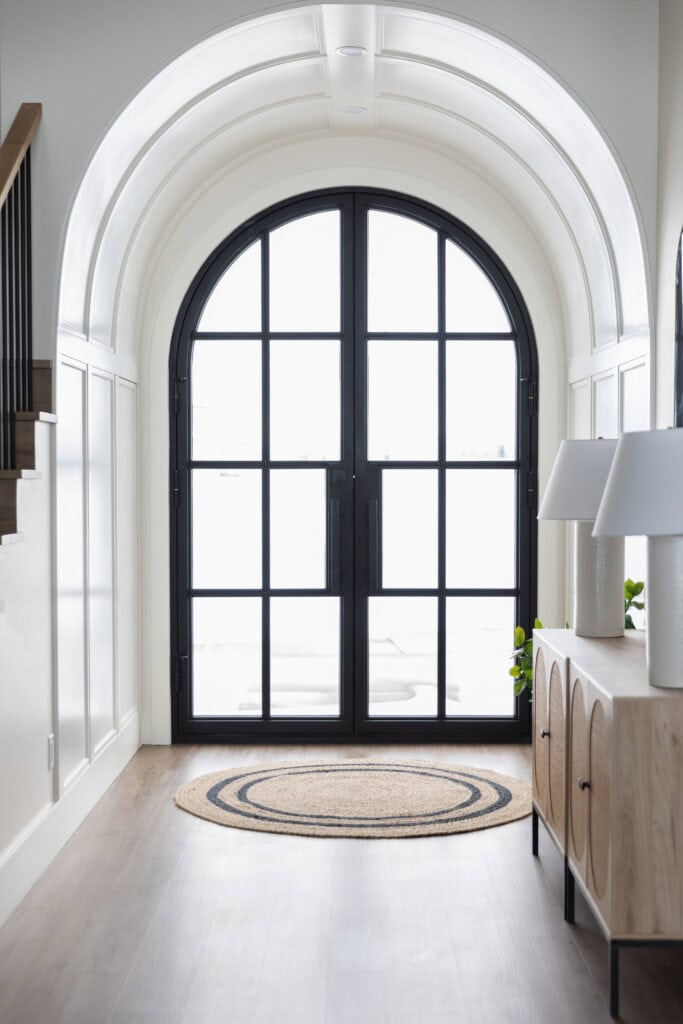 Bright entryway with an arched black-framed glass door, white walls, a round woven rug on wood flooring, and a light wood console table with two white lamps and a small green plant.