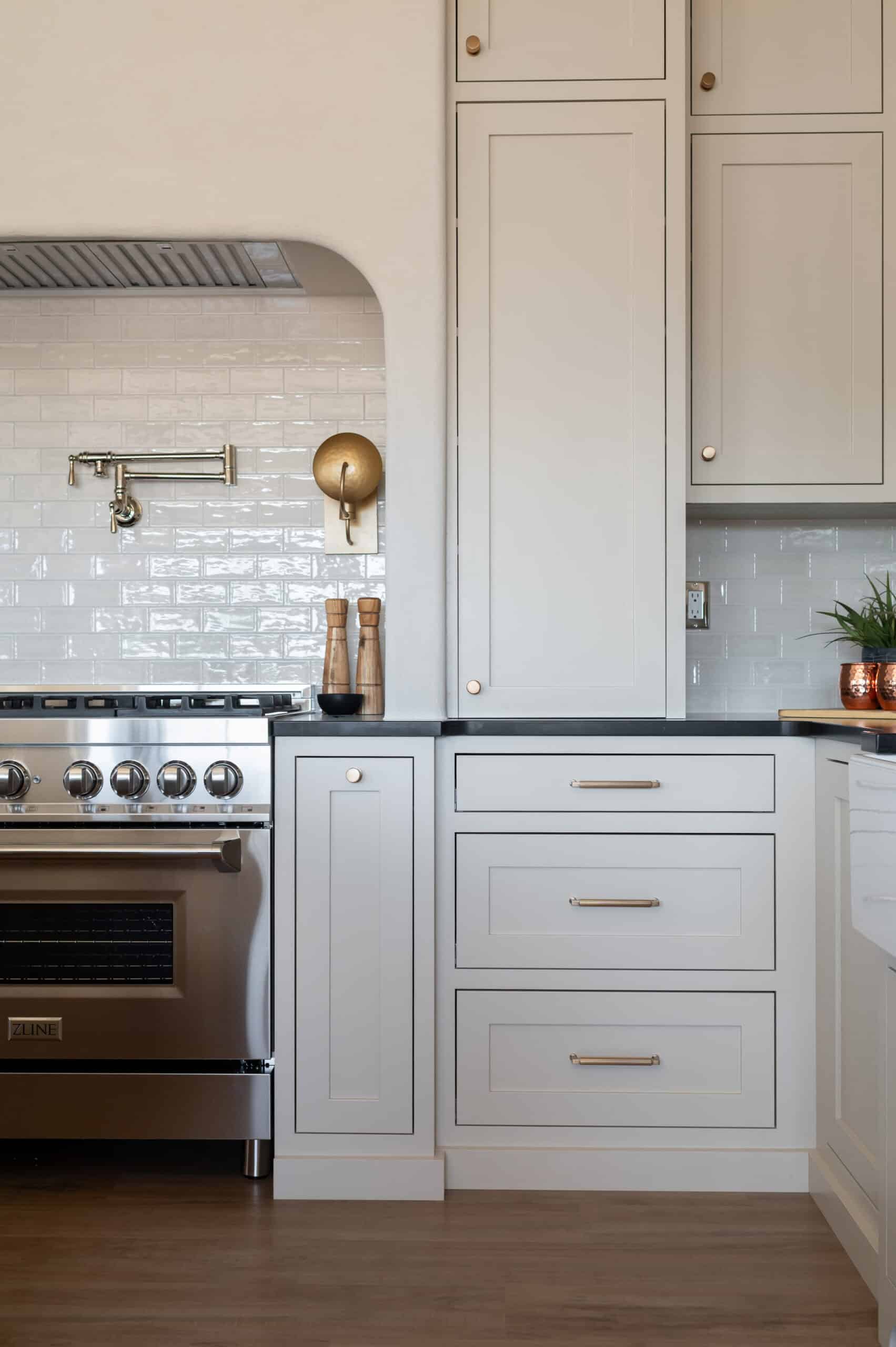 A modern kitchen with light gray cabinets, brushed gold hardware, a stainless steel stove, white subway tile backsplash, a wall sconce, and wooden salt and pepper grinders on a dark countertop.