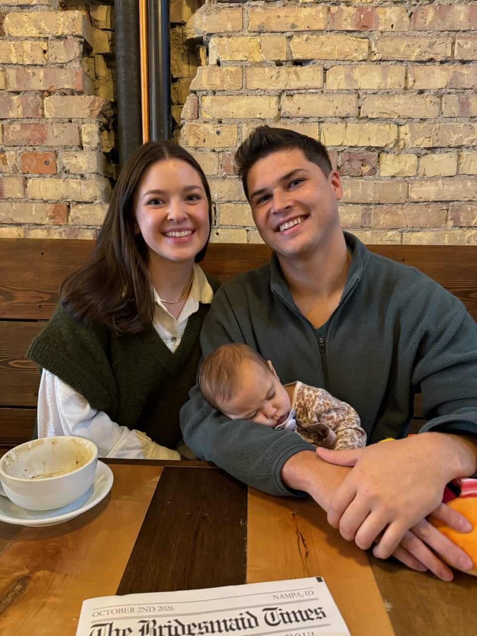 A smiling couple sits at a wooden table in a cozy downtown Twin Falls café, with a baby asleep in the man's arms. An empty coffee cup and a newspaper titled Bridesmaid Times rest on the table against a brick wall background.