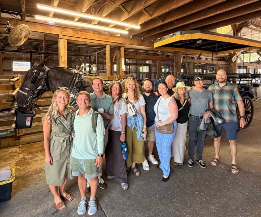 A group of Artisan Builder Collective attendees poses together inside a stable beside a horse and carriage.