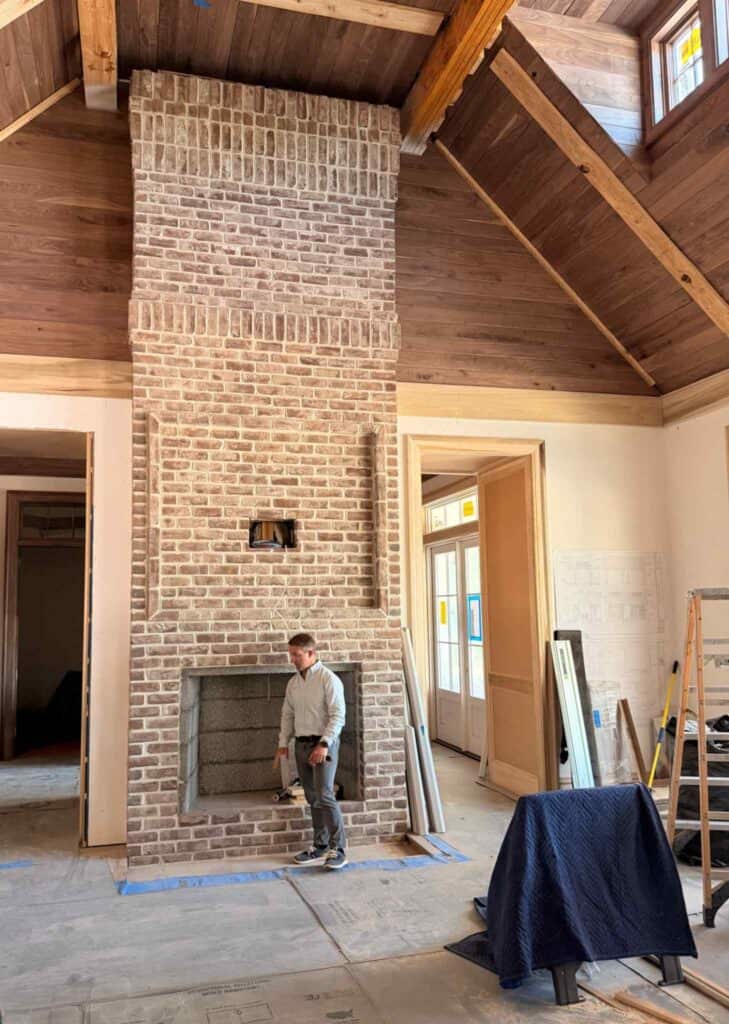 Custom home interior under construction with a tall brick fireplace, vaulted wood ceiling, exposed beams, and building materials throughout the room.
