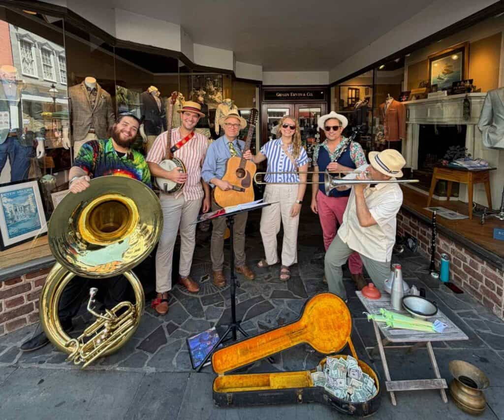 A group of street musicians with brass, string, and percussion instruments performs outside a storefront in Charleston.